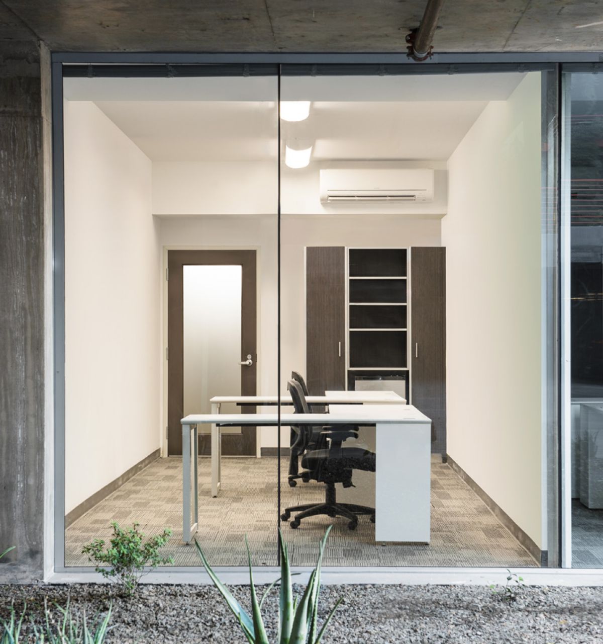 Modern, minimalist office with a glass wall, visible desk, chair, shelves, and small plants outside at Optima Sonoran Village apartments.