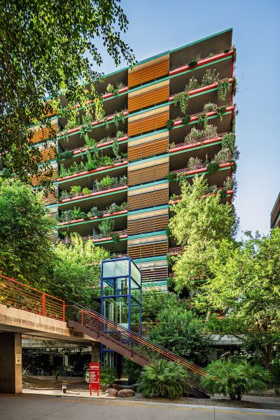 Tall building with colorful horizontal slats and lush greenery on each balcony, surrounded by trees and plants.