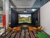 Indoor golf simulator setup with screen, green mat, four orange chairs, and a colorful carpet—perfect for virtual golf sessions.