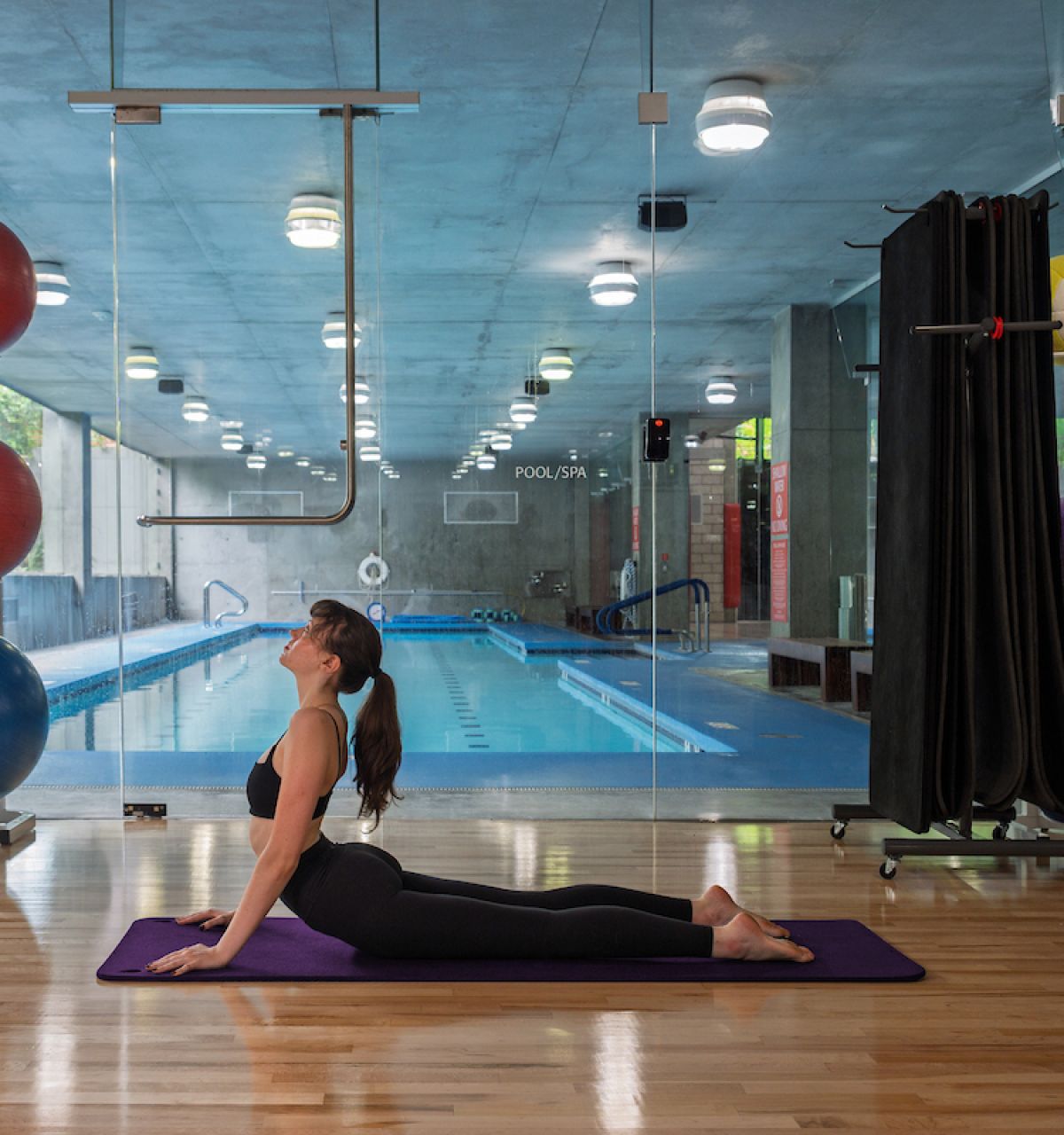 Woman practices yoga in upward-facing dog pose by an indoor pool at Optima Sonoran Village, with Pilates balls nearby.