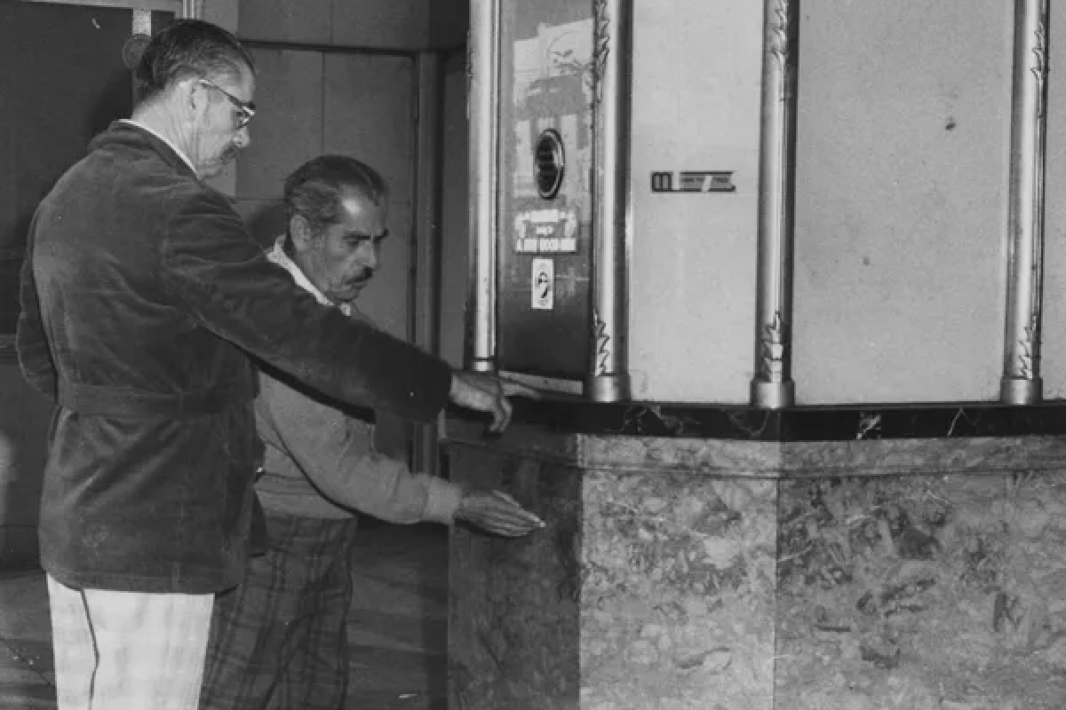 Two men stand at an ornate marble Phoenix theater ticket booth, one gesturing towards the counter.