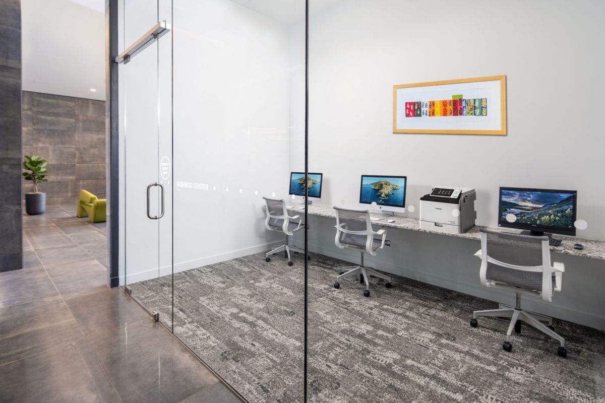 A modern business center by Optima Communities with computers, chairs, printer, and artwork—ideal for hybrid working behind glass doors.