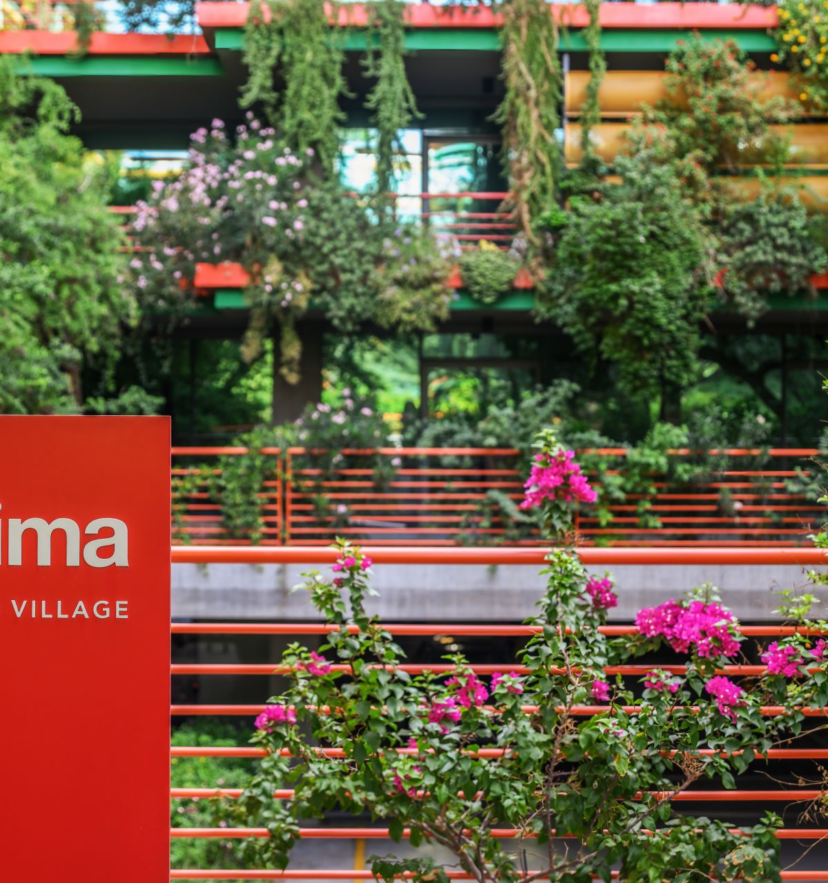 Red "Optima Sonoran Village" sign in front of lush greenery and modern building showcasing sustainable architecture with plants and flowers.