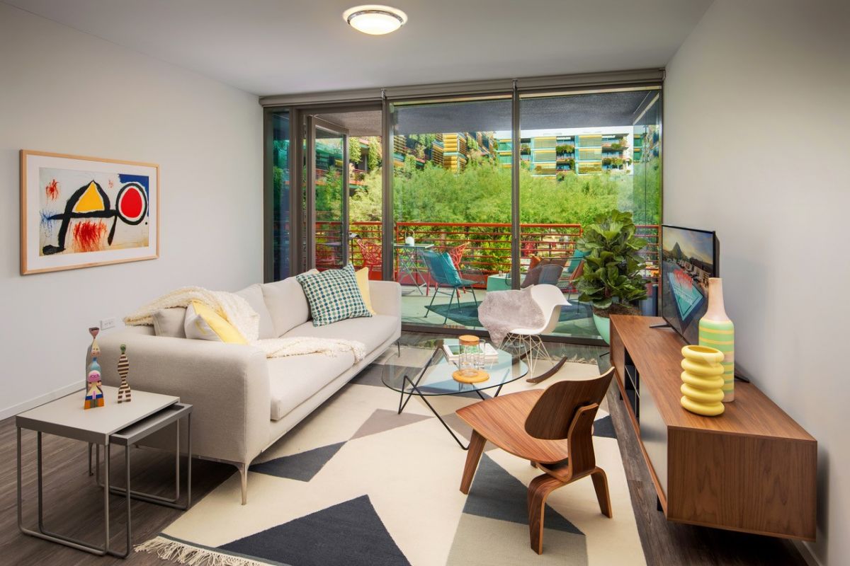 Modern apartment living room with a cream sofa, wooden furniture, art pieces, and a balcony overlooking lush greenery.