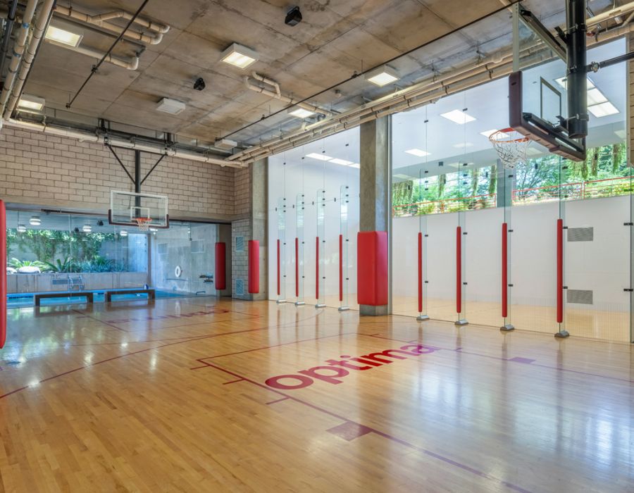 Indoor basketball court with glass walls, a wooden floor, nets, and padded red accents at Optima Sonoran Village apartments.