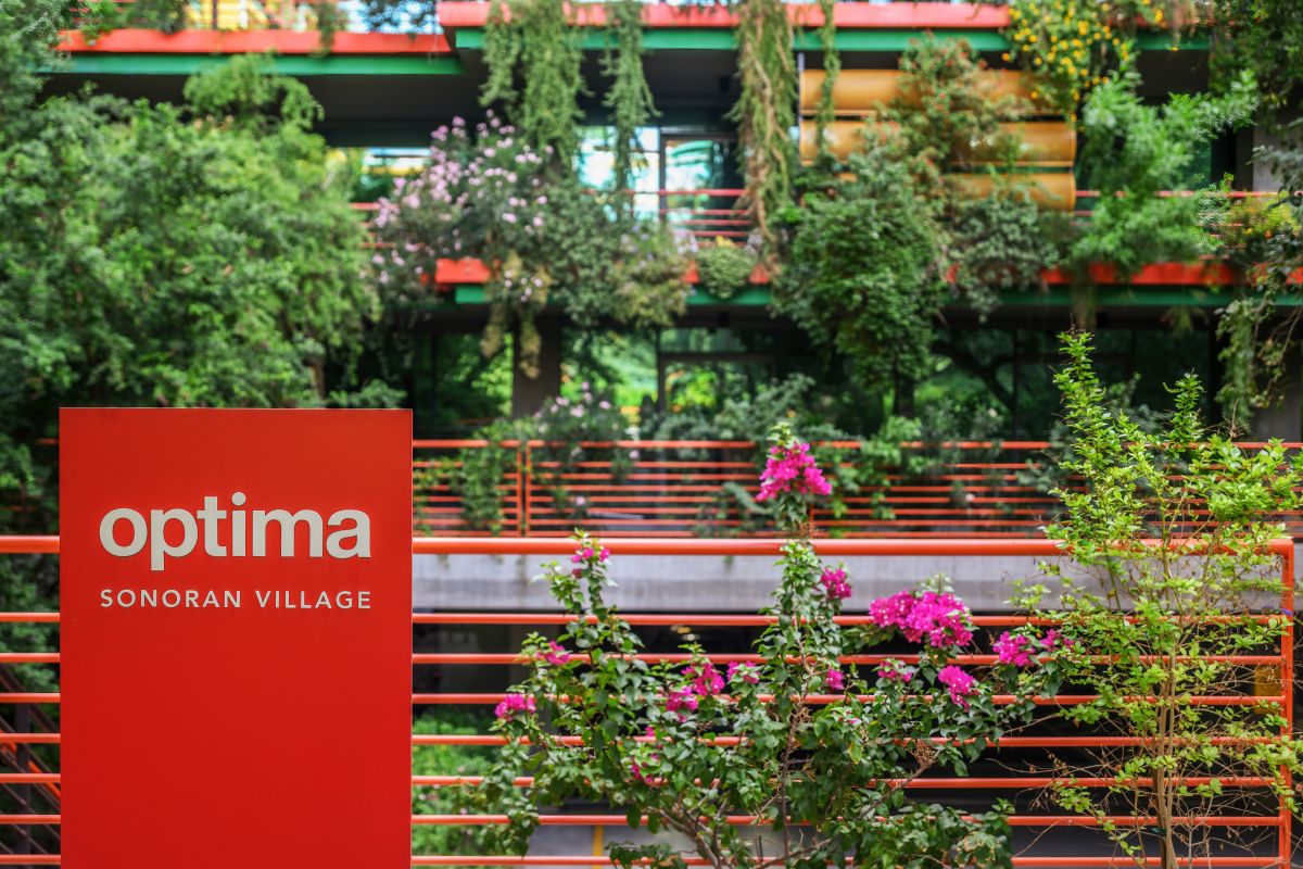Red "Optima Sonoran Village" sign in front of lush greenery and modern building showcasing sustainable architecture with plants and flowers.