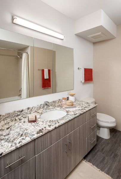 Modern bathroom with a granite countertop, large mirror, wood cabinetry, and red towel accents at Optima Sonoran Village apartments.