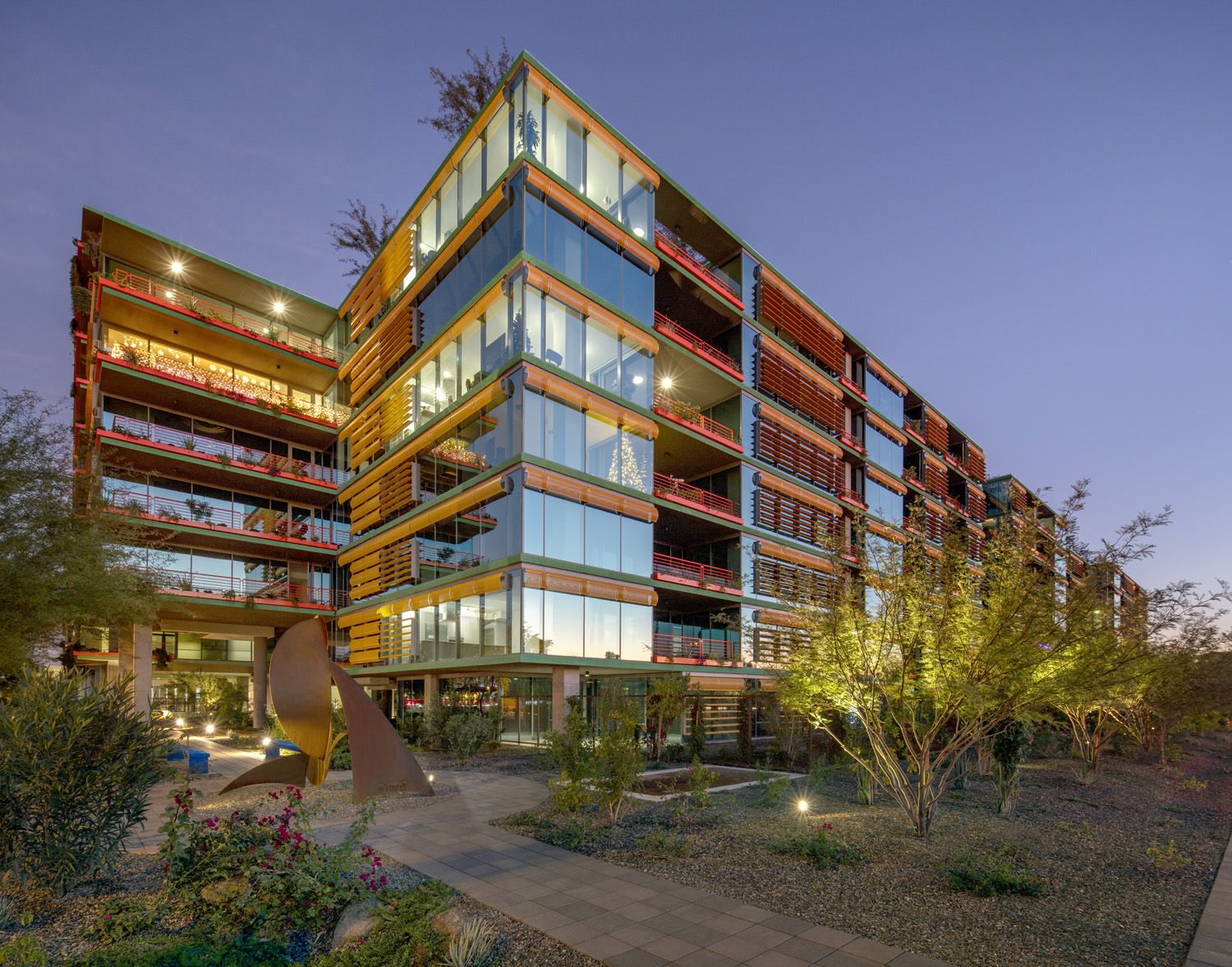 Modern glass and steel apartment at dusk, like Optima Sonoran Village, surrounded by greenery and illuminated lights.