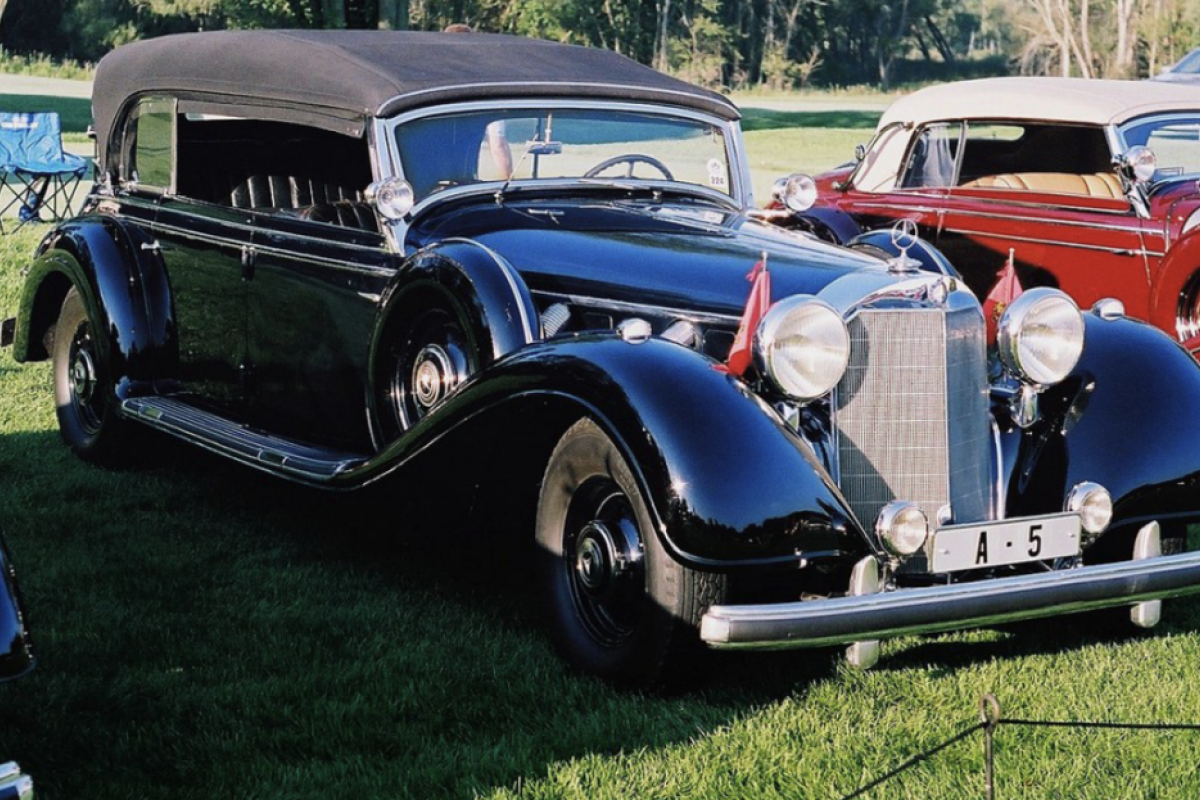 A shiny black vintage Mercedes-Benz convertible is parked on grass at a classic car show, reminiscent of Scottsdale's Barrett-Jackson event.