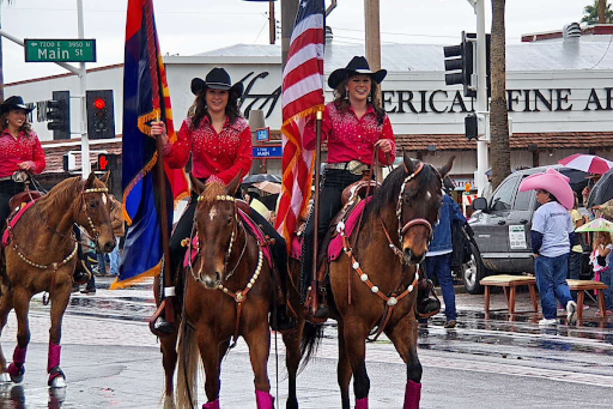 Two women in red shirts ride horses in a parade during Scottsdale Western Week, carrying American and state flags on a rainy street.