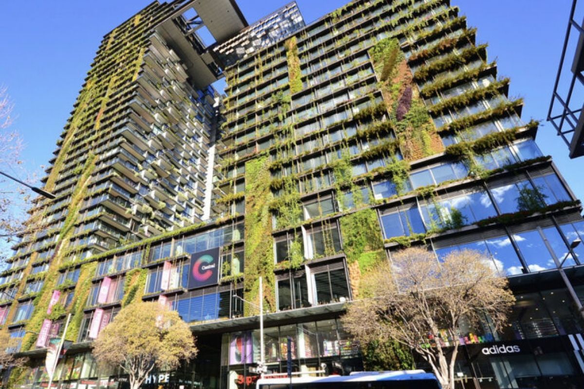 Tall modern building featuring green walls and vertical landscaping, with shops and cars bustling at street level.