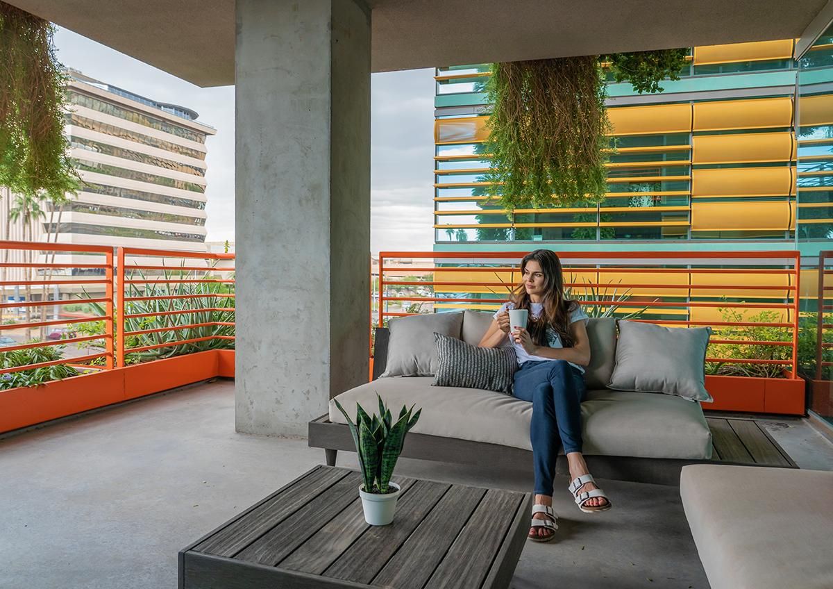 Woman sitting on outdoor couch, holding a drink, surrounded by modern architecture and greenery at Optima Sonoran Village apartments.