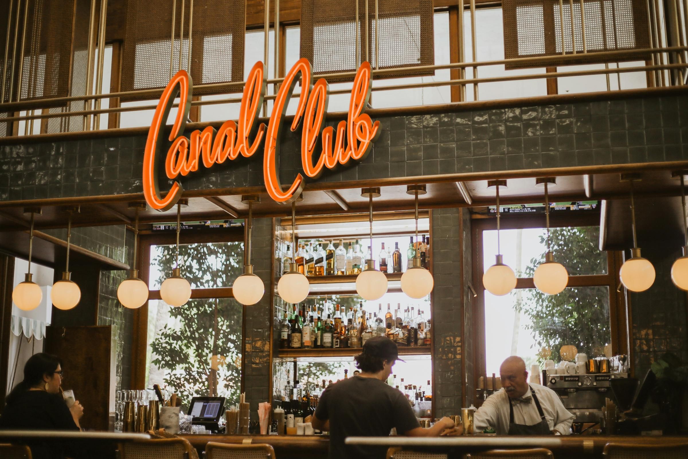 A bartender serves a customer at a stylish bar with a neon "Canal Club" sign and shelves of bottles behind them in Scottsdale, near Optima Sonoran Village apartments.