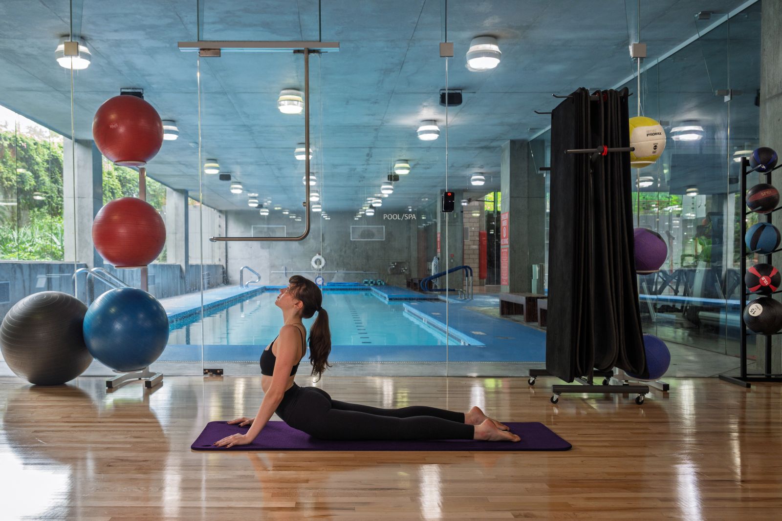 Woman doing yoga in upward-facing dog pose on a mat, beside a pool in a modern gym.