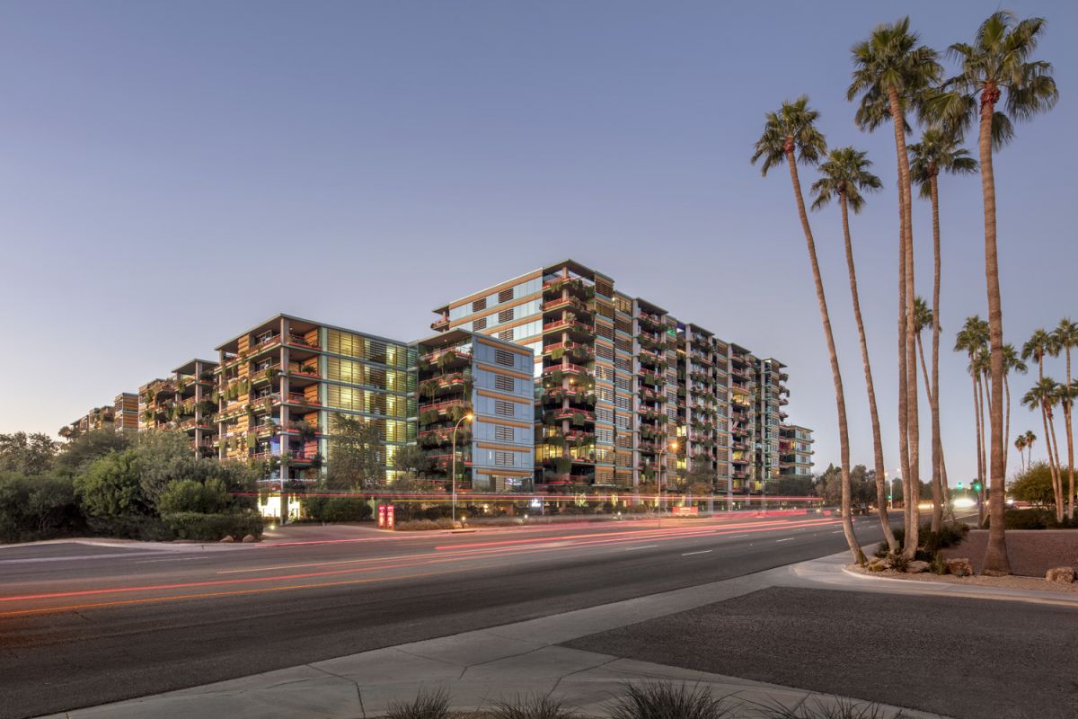 Modern multi-story apartment buildings at dusk, with palm trees and light trails from passing cars in the foreground.
