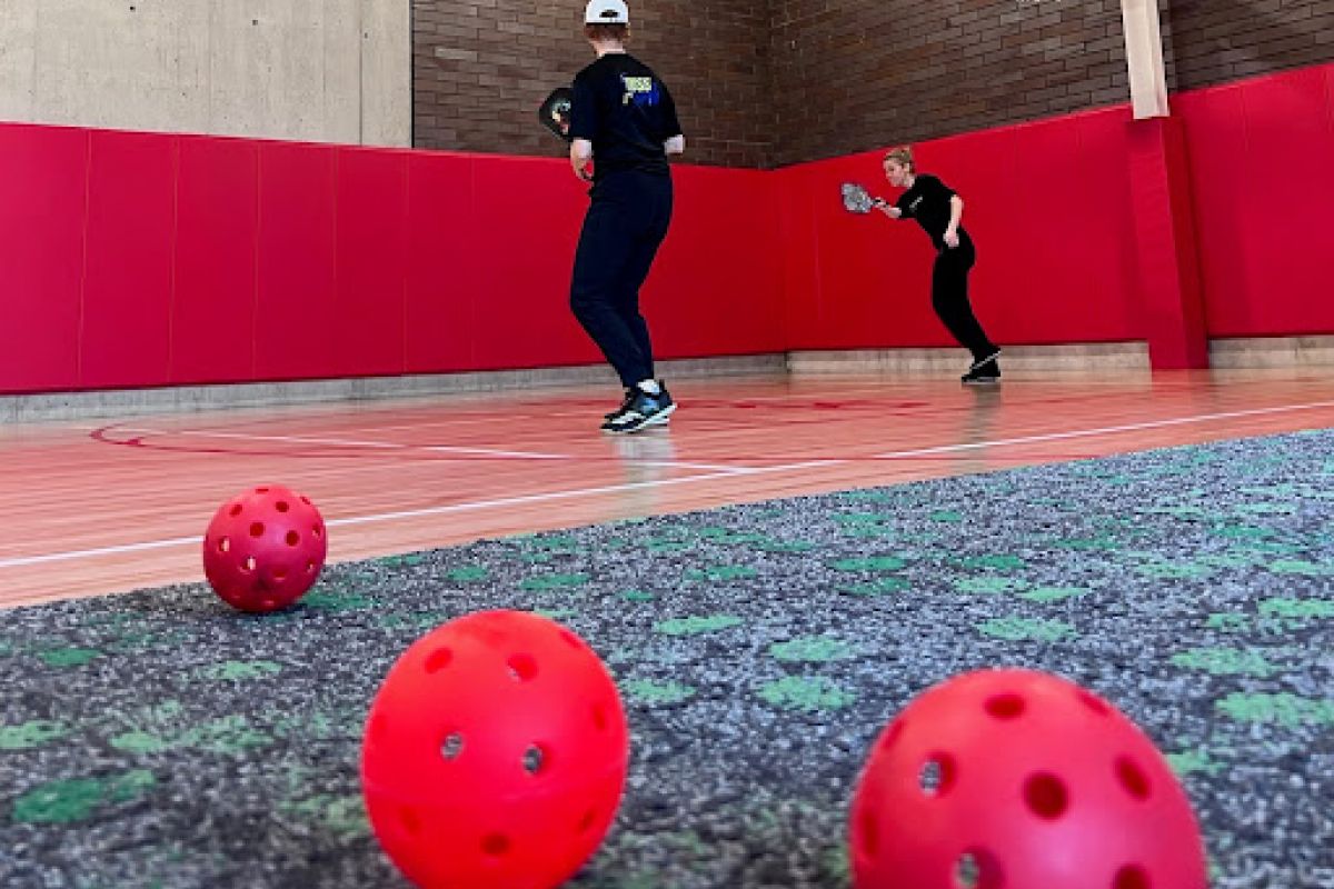 Two people play pickleball in a gym, fostering connection as red perforated balls are scattered on the floor in the foreground.