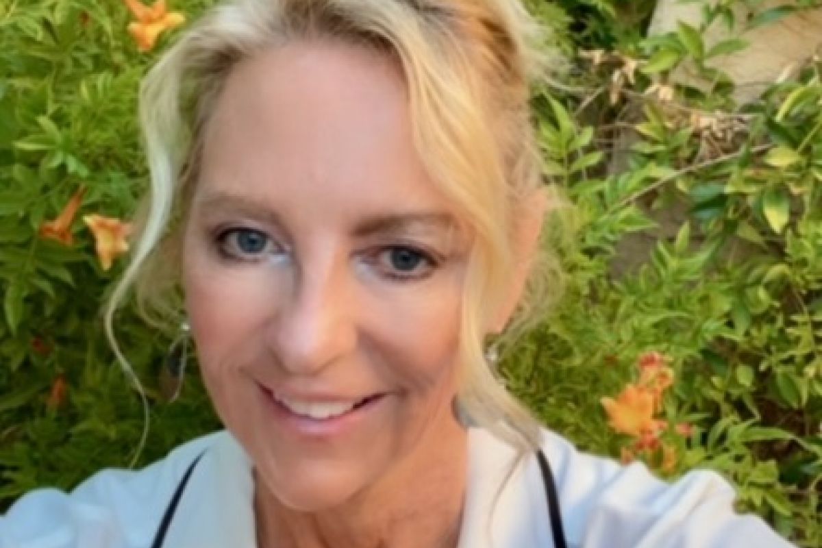 Woman with blonde hair and white top smiling outdoors at Optima Sonoran Village in front of green plants with orange flowers.