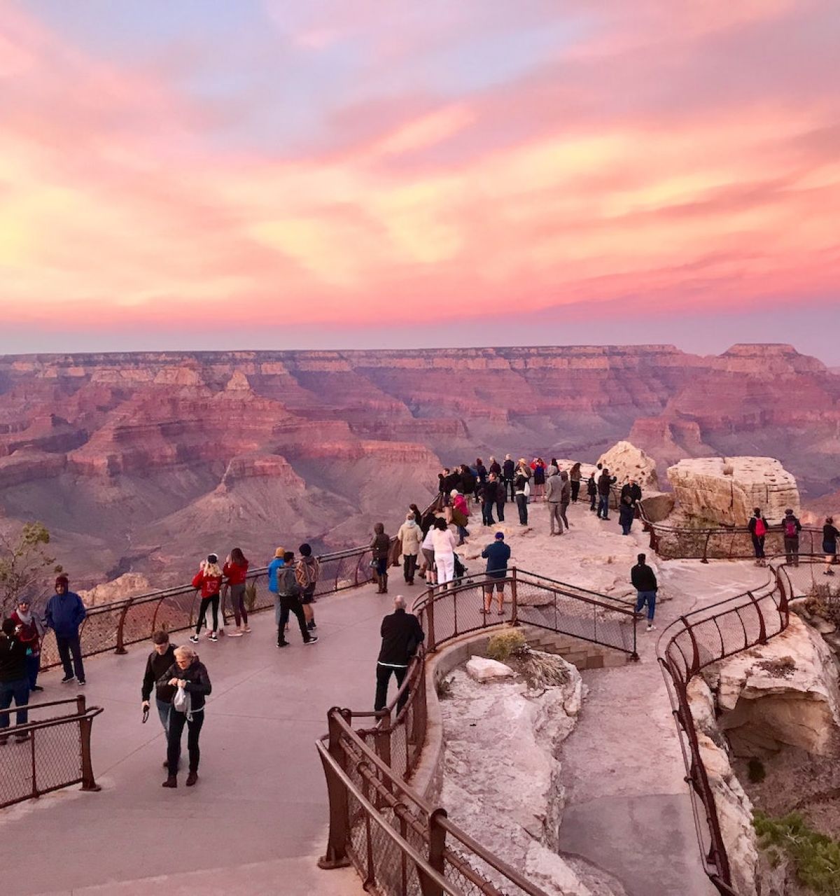 People gather at a Grand Canyon viewpoint at sunset, a favorite for day trips from Phoenix and Scottsdale under a colorful pink and orange sky.