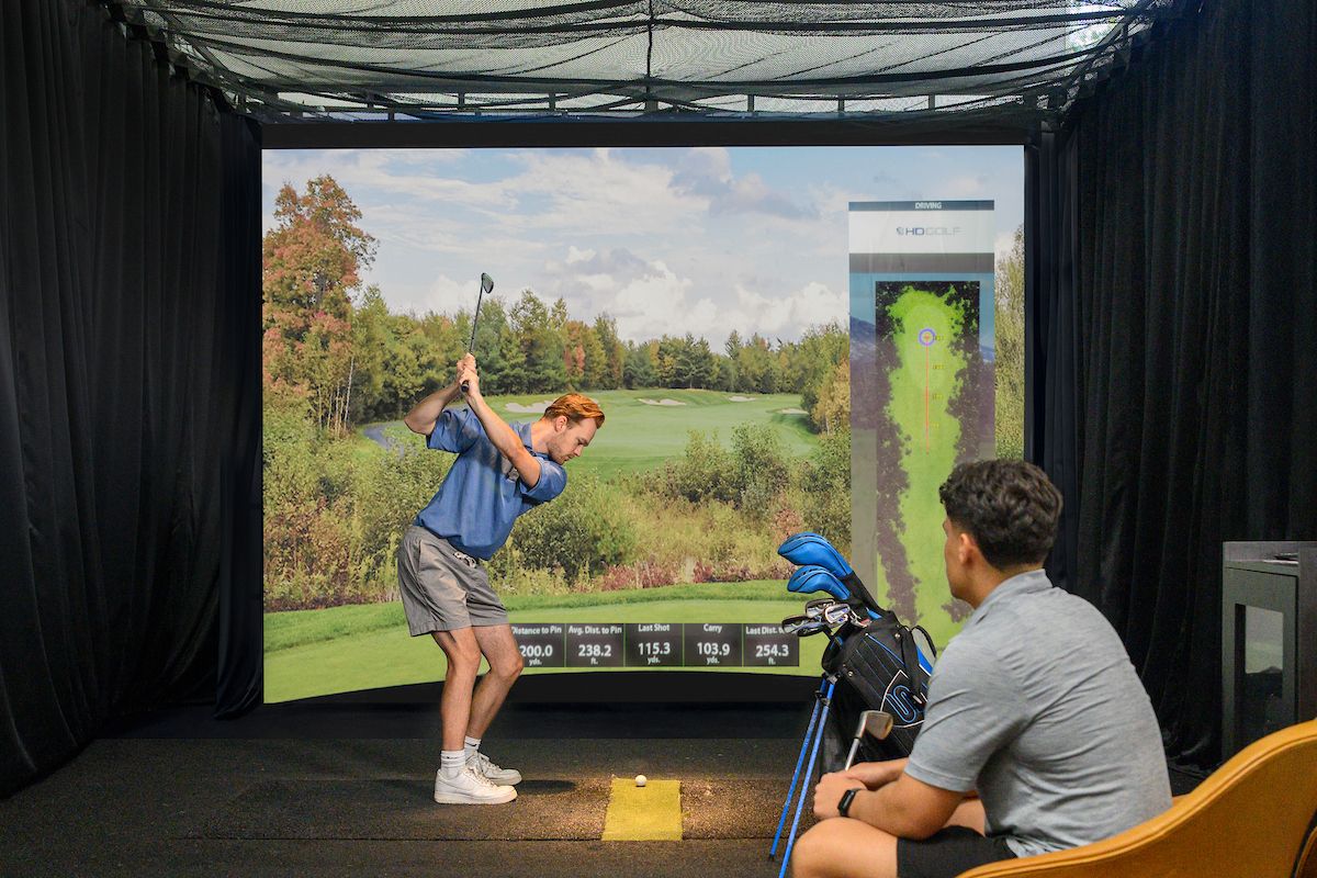 Two people enjoy daily life at Optima Community, using an indoor golf simulator—one swings while the other watches by the golf bag.