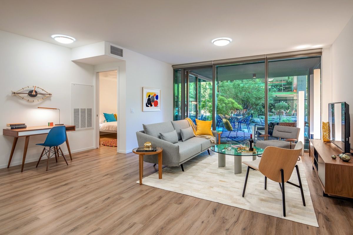 Modern living room in a 2-3-bedroom apartment with wood floors, stylish furniture, and large windows opening to a patio with greenery.