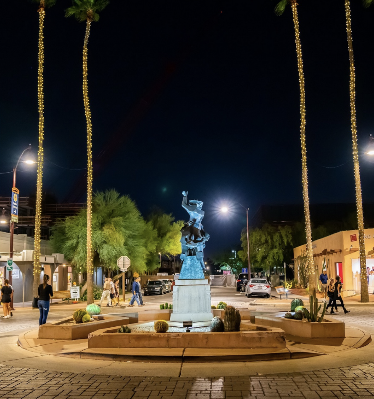 Nighttime street scene with a bronze statue in a roundabout, glowing palm trees, and Scottsdale ArtWalk ambiance.