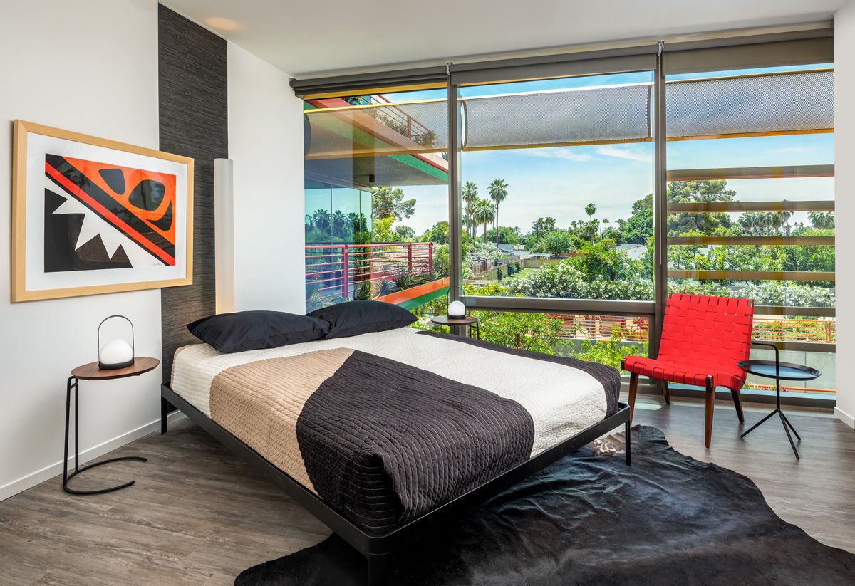 Modern bedroom with large windows, double bed, abstract art, red chair, and views of trees and sky at Optima Sonoran Village apartments.