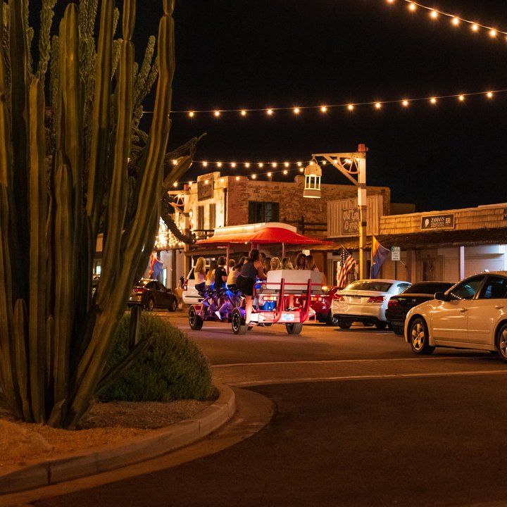 Street at night in Downtown Scottsdale with string lights, a cactus, parked cars, and a group riding a pedal bar with red canopy.