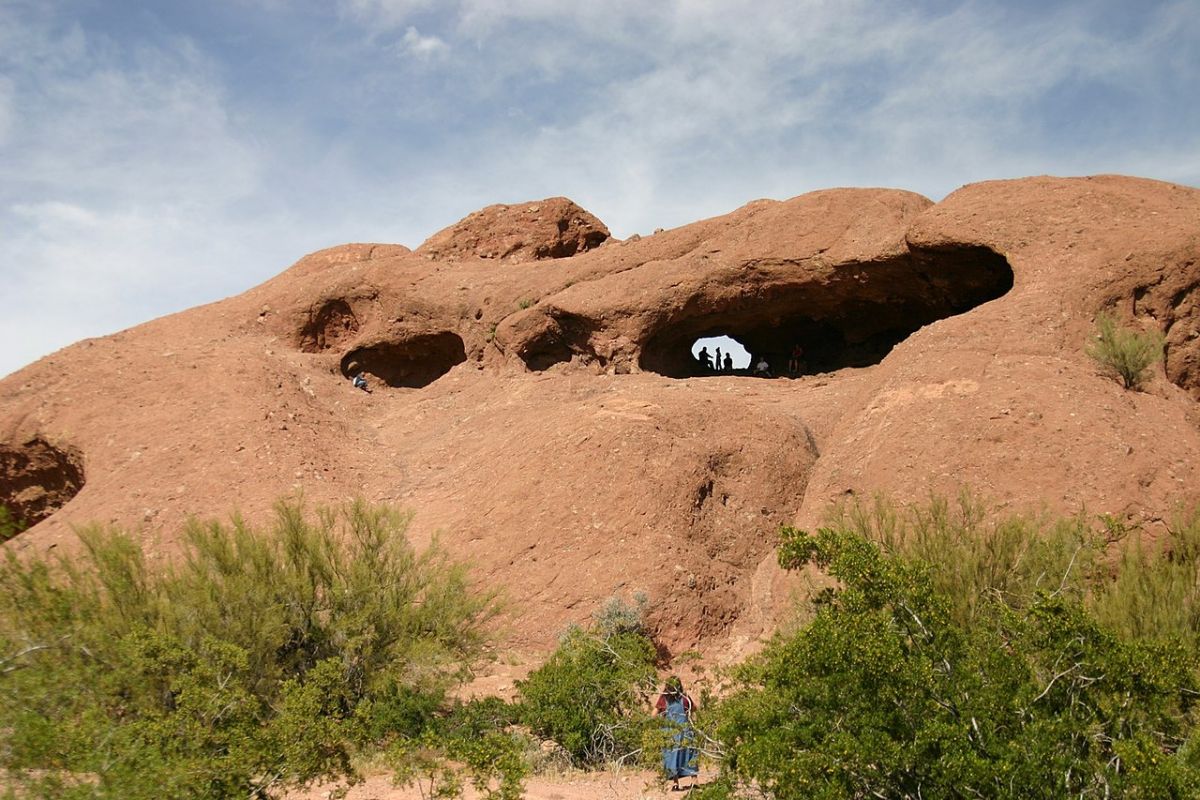 Large red rock formation with natural holes at Papago Park; people silhouetted inside, greenery and hiking trails in front.