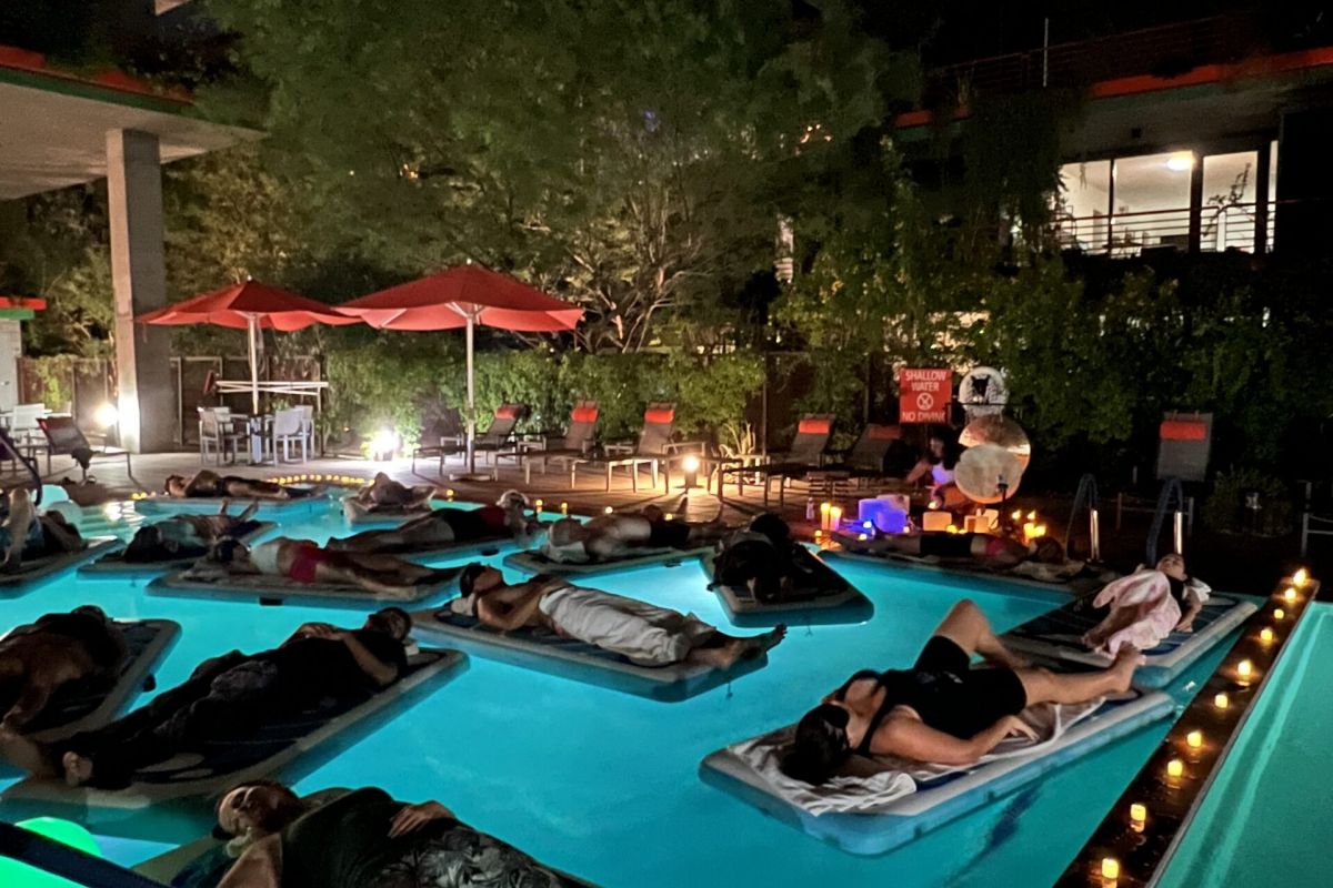 People relaxing on floating mats in a pool at night during Optima Communities' celebration events, surrounded by candles and red umbrellas.