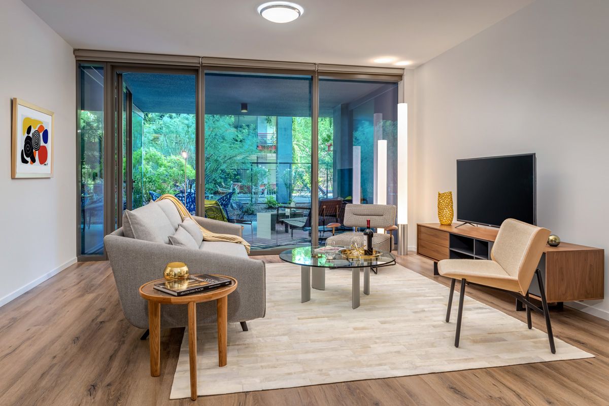 Optima Sonoran Village modern living room with large windows, gray sofa, beige chairs, glass coffee table, and wooden flooring.