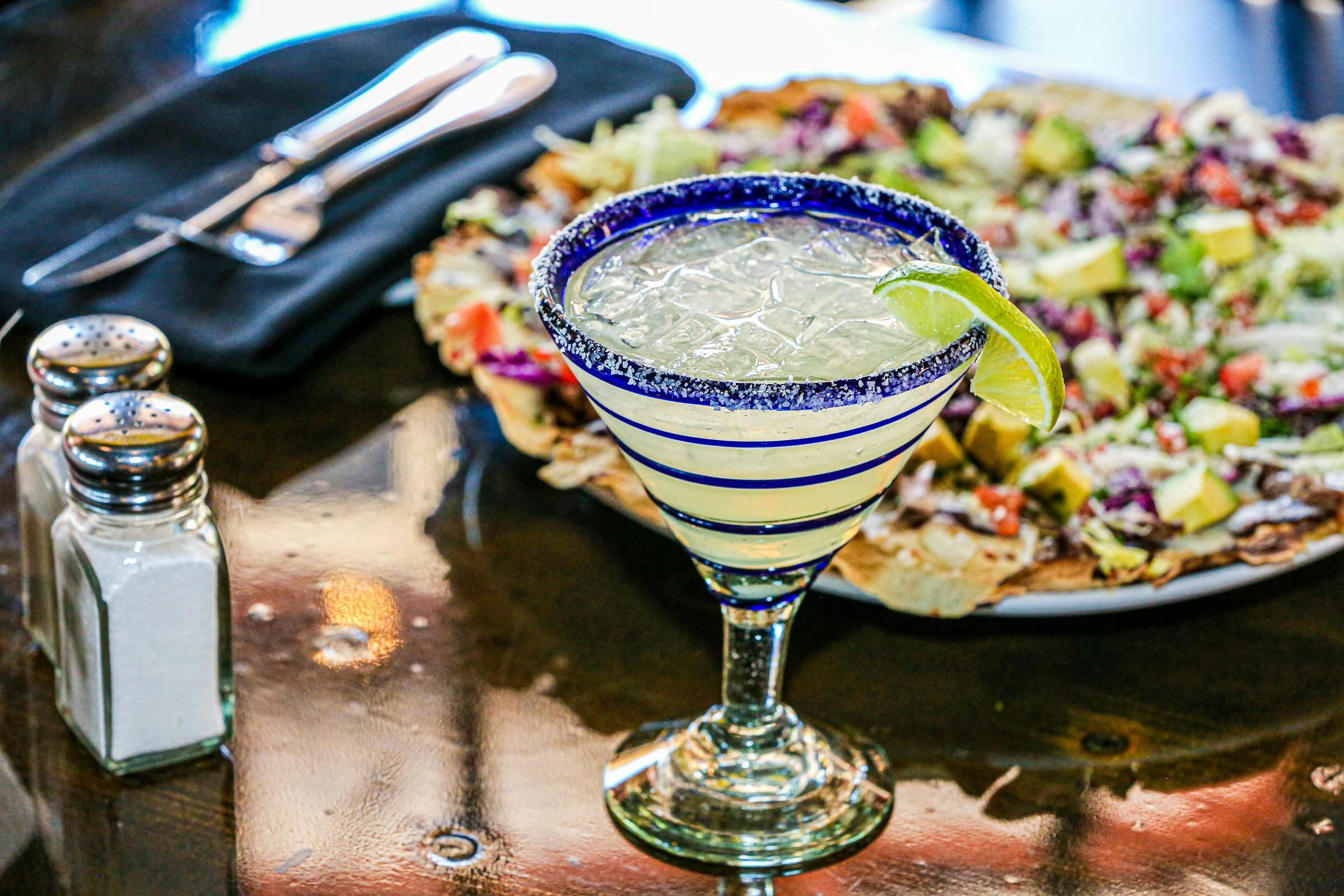 A margarita with a lime wedge on a table, next to salt and pepper shakers, with food blurred in the background.