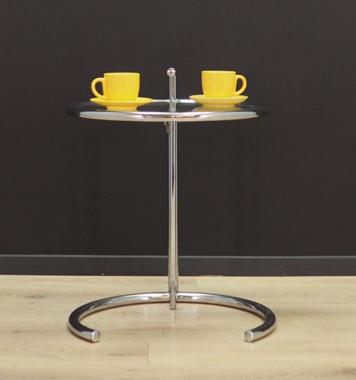 A spotlight shines on a round chrome Table E-1027 with two yellow cups, set against a black wall and wooden floor.