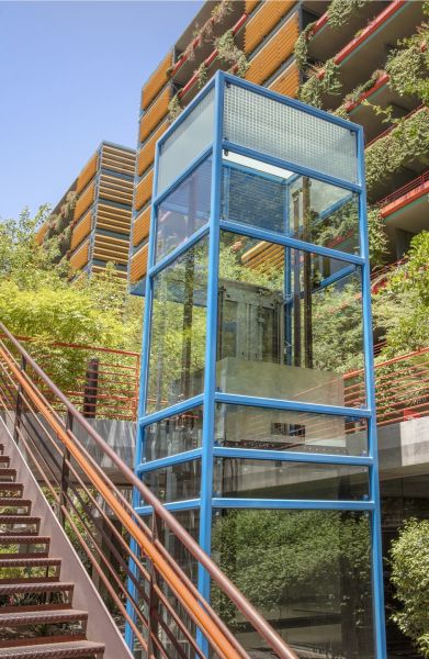 Optima Sonoran Village apartments glass elevator with a blue frame next to stairs, surrounded by greenery and a colorful building facade.