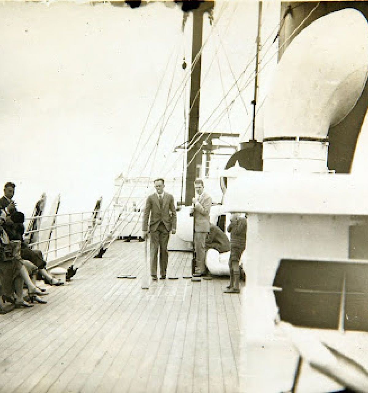 People on the deck of a ship; some seated, some standing, gathering in a community around ship equipment in the foreground.