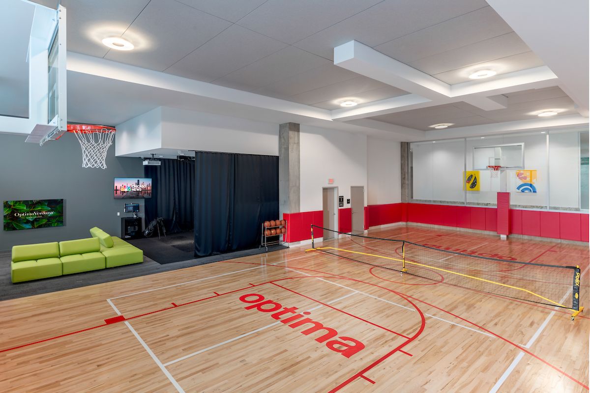 Indoor gym with a basketball hoop, mini tennis net, red markings, and a green couch near a TV in the corner.