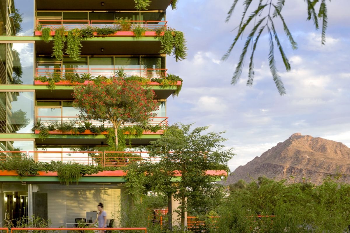 Modern apartment building with lush balcony plants, mountain in background, and person walking below.