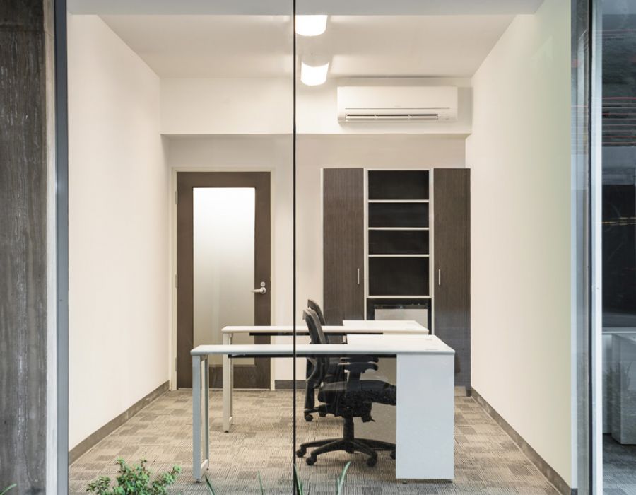 Modern, minimalist office with a glass wall, visible desk, chair, shelves, and small plants outside at Optima Sonoran Village apartments.