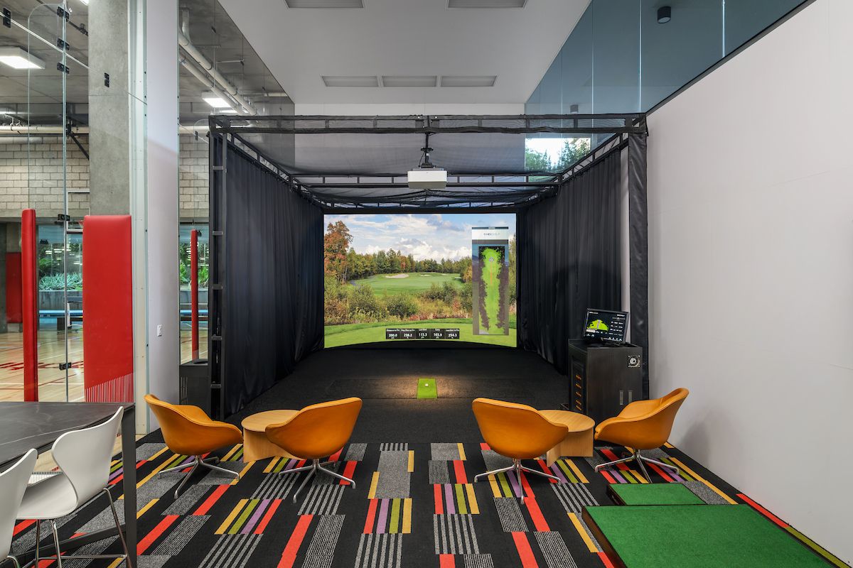 Indoor golf simulator setup with screen, green mat, four orange chairs, and a colorful carpet—perfect for virtual golf sessions.