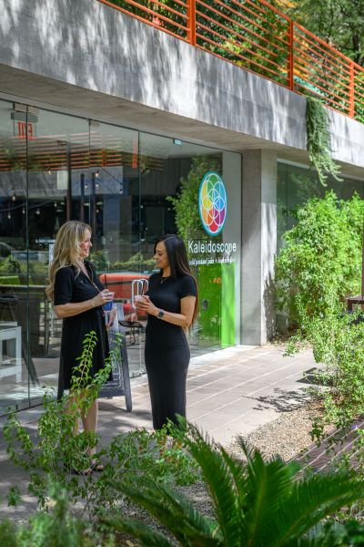 Woman in a floral dress stands by a glass door labeled "Kaleidoscope;" greenery surrounds the entrance at Optima Sonoran Village apartments.