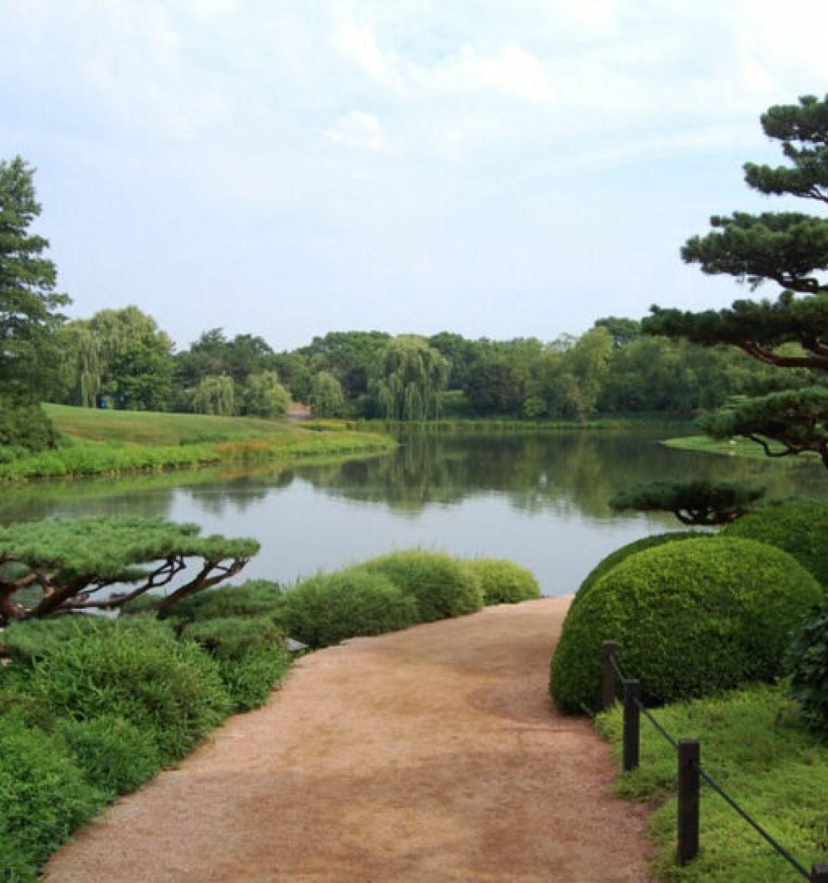 Pathway in a Japanese garden beside a calm pond, surrounded by sculpted trees and lush greenery—perfect for serene autumn travel.