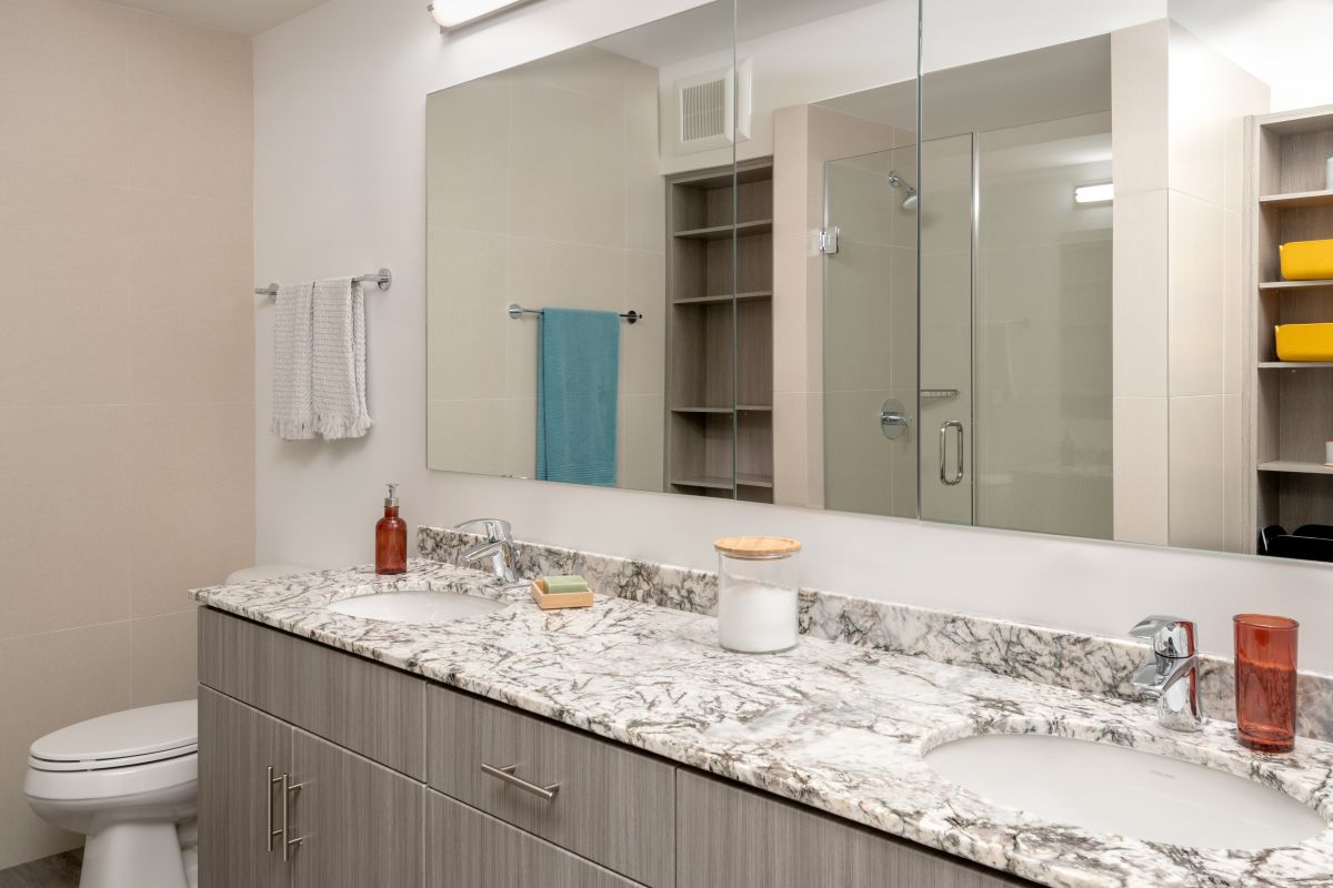 Modern bathroom design with a marble countertop, double sinks, large mirror, toilet, and wooden cabinets under bright lighting.
