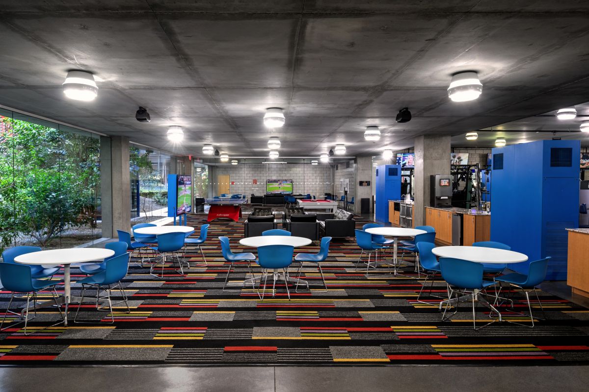 Modern lounge with blue chairs, round tables, colorful carpet, vending machines, and large windows overlooking greenery.