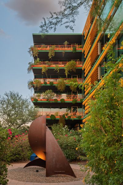 A modern building with lush plants on balconies and a rust-colored abstract sculpture in front.