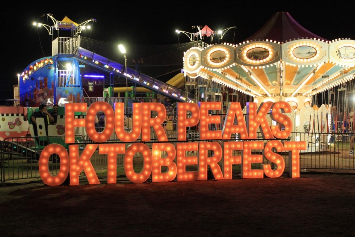 A glowing "Four Peaks Oktoberfest" sign shines at Tempe Beach Park, set against the backdrop of lively carnival rides at night.
