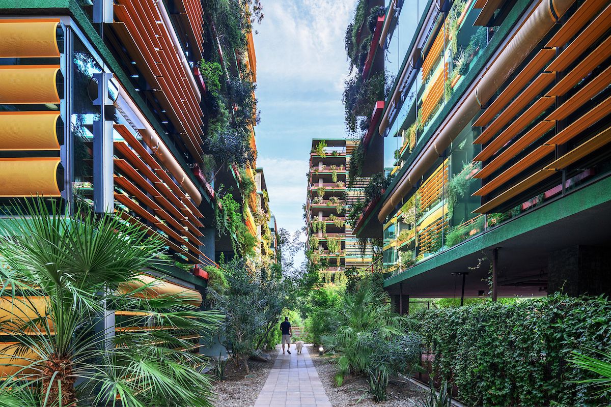Two people walk between pet-friendly, modern green buildings at Optima Sonoran Village, surrounded by orange sunshades and lush plants.