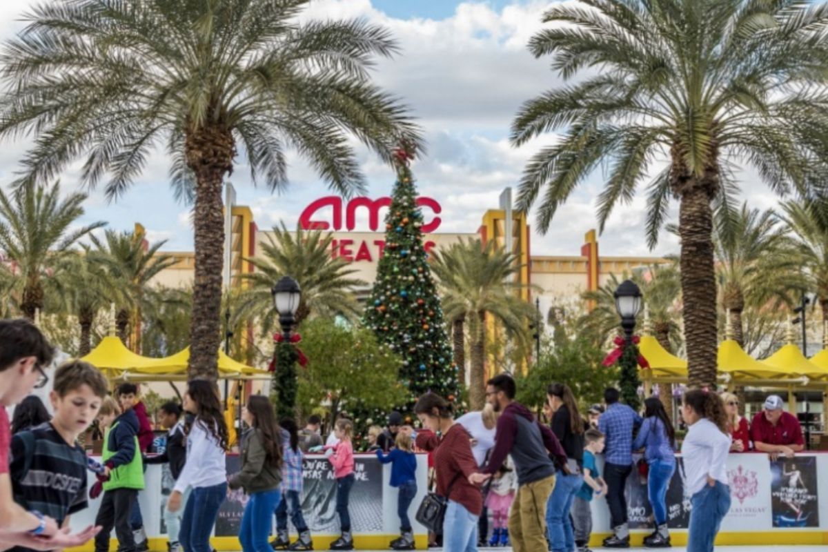 People ice skating outdoors near a Christmas tree and palm trees—Optima’s Guide to Thanksgiving 2024 fun by the AMC Theatres sign.