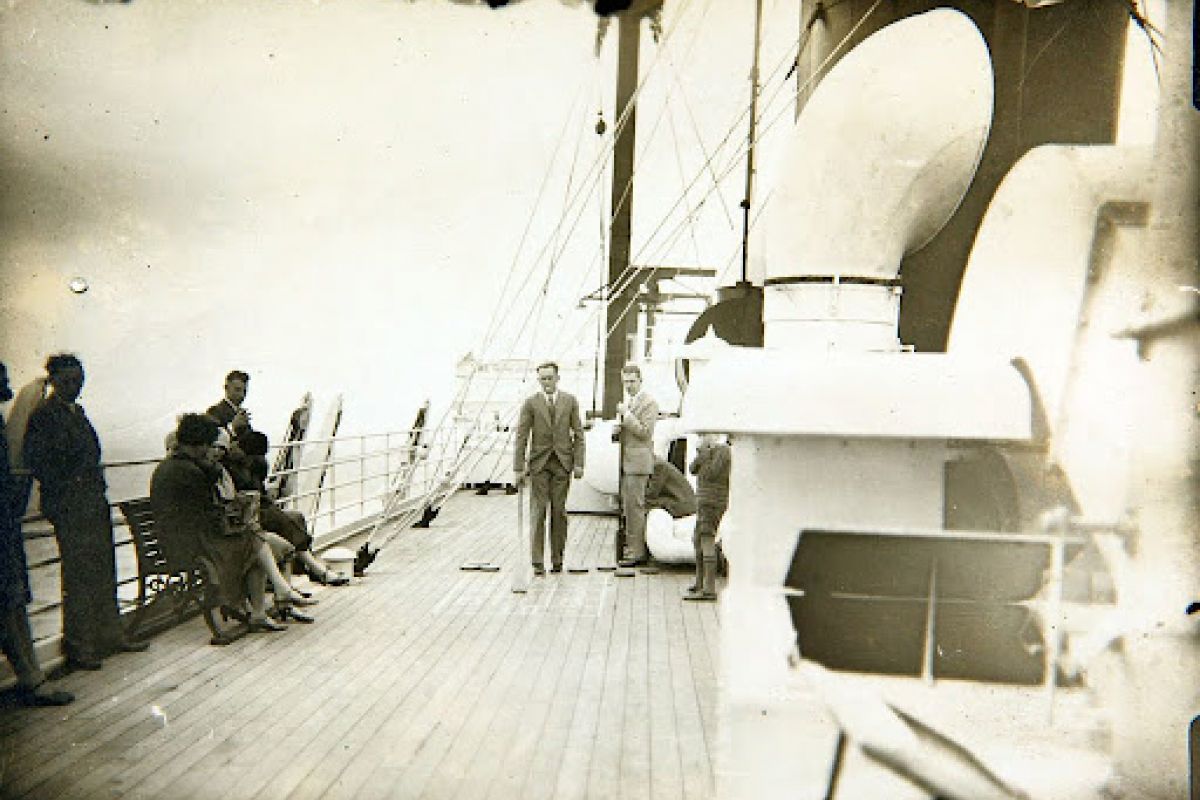 People on the deck of a ship; some seated, some standing, gathering in a community around ship equipment in the foreground.