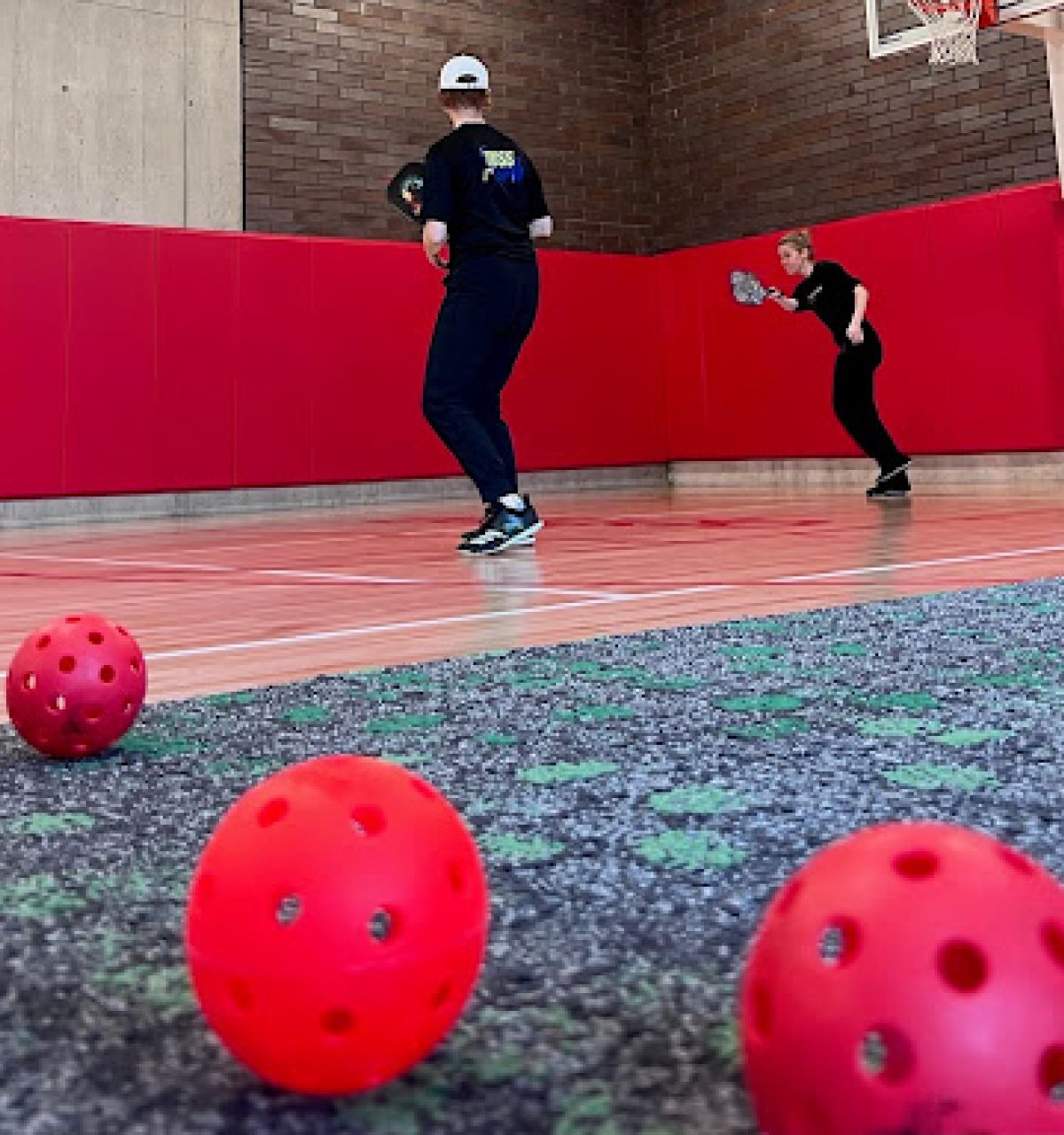 Two people play pickleball in a gym, fostering connection as red perforated balls are scattered on the floor in the foreground.