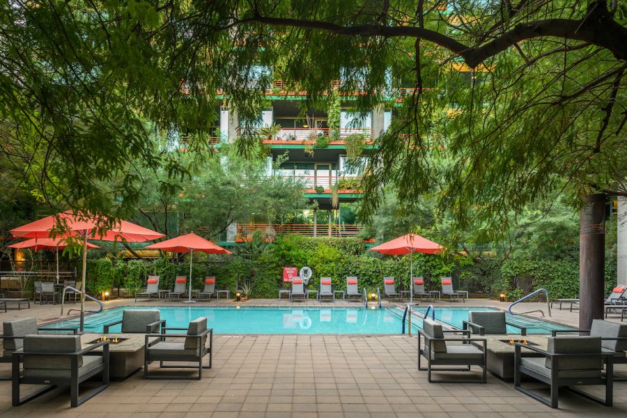 Outdoor pool surrounded by lounge chairs, red umbrellas, lush trees, and a modern building in the background.