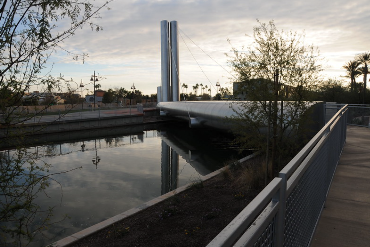 Modern Soleri Bridge with tall silver pillars spans a canal in Scottsdale, surrounded by plants and glowing at sunset.